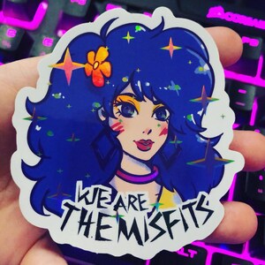 May include: A sticker featuring a cartoon woman with blue hair, a yellow flower, and sparkling stars. The text "WE ARE THE MISFITS" is printed in black on the sticker.