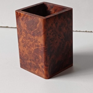 May include: A small, square wooden container with a natural wood grain pattern. The container is made of a reddish-brown wood with a light brown interior.