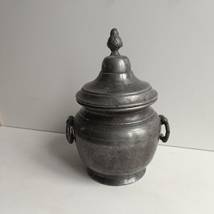 Puede incluir: Un pequeño recipiente metálico antiguo con tapa. El recipiente es de color gris oscuro con una superficie texturizada y dos asas pequeñas. La tapa tiene un remate decorativo. El recipiente es probablemente para almacenamiento.