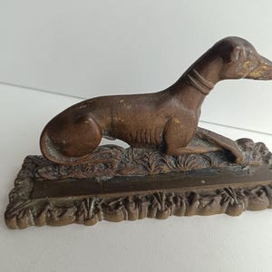 Könnte beinhalten: Eine bronzefarbene Hunde-Figur in Ruheposition. Der Hund hat einen schlanken Körper und einen langen Hals mit Halsband. Er ruht auf einem rechteckigen Sockel mit einem verzierten, strukturierten Rand. Die Figur hat ein Vintage-Aussehen.