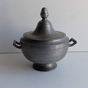 Puede incluir: Una sopera de peltre plateado con tapa y dos asas. La sopera tiene un cuerpo redondo y una tapa abovedada con un pomo.