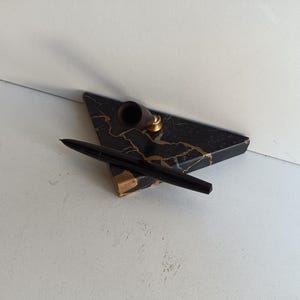 Op de afbeelding: Een zwarte pen en penhouder set. De penhouder heeft een driehoekige vorm met een zwart en goud marmerpatroon. De pen is zwart en rust op de houder. De penhouder heeft een kleine messingkleurige ring.
