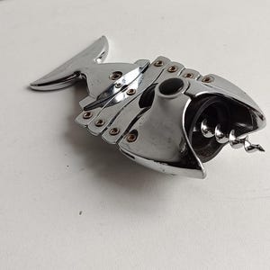 May include: A silver fish-shaped bottle opener with a black handle and a corkscrew. The opener has a hinged design and a series of small metal rings.