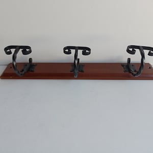May include: A wooden coat rack with three decorative black metal hooks. The hooks have a scrolled design and are mounted on a rectangular, dark brown wooden board. The rack is designed for wall mounting.