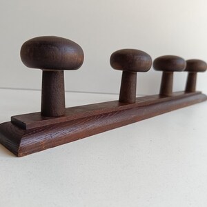 May include: A dark brown wooden coat rack with four mushroom-shaped pegs. The pegs are attached to a rectangular base. The wood grain is visible, and the rack is set against a neutral background.