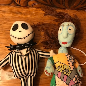 May include: A pair of plush toys from the Nightmare Before Christmas movie. The first toy is a black and white striped Jack Skellington with a black bow tie. The second toy is a blue and green Sally with brown hair.