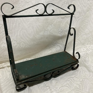 Puede incluir: Estantería de metal verde oscuro con base rectangular y parte superior decorativa con volutas. La estantería tiene un acabado envejecido, lo que sugiere un estilo antiguo o vintage. El marco es de hierro forjado.