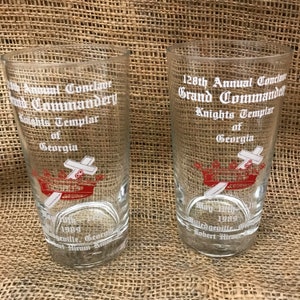 May include: Two clear glass tumblers with white lettering. The tumblers commemorate the 128th Annual Conclave Grand Commandery Knights Templar of Georgia. The tumblers feature a red and white crown with a sword and cross.