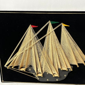 Peut inclure: Une décoration murale en art de fil représentant un voilier à trois voiles. Les voiles sont faites de fil doré et sont tendues entre des clous sur un fond de velours noir. Le voilier a trois petits drapeaux, un rouge, un vert et un jaune.