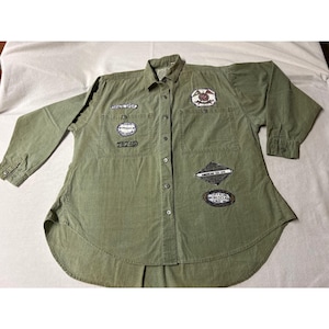 Puede incluir: Camisa verde oliva con botones y múltiples parches. Los parches incluyen texto y diseños gráficos. La camisa tiene mangas largas y dos bolsillos en el pecho. La camisa tiene un dobladillo curvo y un cuello.