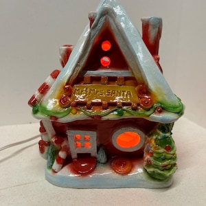 May include: A red and white ceramic Christmas house with a lit window and the words "Mr. & Mrs. Santa" on the front. The house has a candy cane trim and a green tree with red ornaments.