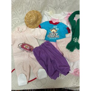 May include: A collection of doll clothing and accessories including a straw hat, a light pink patterned onesie with white foot covers, a bright blue t-shirt with a monkey graphic and text, purple pants, a green scarf with a snowflake, and a pink toy.