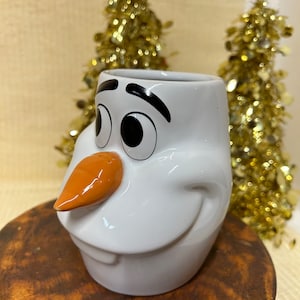 May include: White ceramic mug shaped like Olaf, the snowman from the movie Frozen. Olaf has a black smile, black eyes, and a black eyebrow. Olaf's nose is orange.