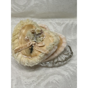 May include: A heart-shaped antique powder puff with a light peach satin top, adorned with a velvet ribbon, lace trim, and small floral accents. The puff sits atop a clear, scalloped glass dish. The overall color palette is soft and vintage.