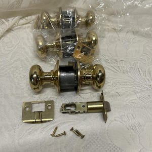 May include: A set of brass-colored door knobs, a strike plate, and a latch bolt. The door knobs are oval-shaped and come with screws for installation. The hardware is new and ready for use.