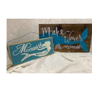 May include: Two rectangular wooden signs. The smaller sign is teal with white text reading "Mermaids Welcome" and a mermaid silhouette. The larger sign is dark brown with a teal whale tail and white text reading "Make Waves little mermaid".