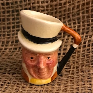 Może przedstawiać: Mały ceramiczny kubek w kształcie głowy mężczyzny w cylindrze. Kubek jest brązowy, biały i żółty z czarną wstążką na kapeluszu. Mężczyzna ma brązowy wąs i brodę.