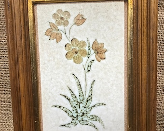 May include: A framed ceramic tile with a floral design. The tile features a stylized plant with brown flowers and green leaves on a white background. The frame is brown wood with gold accents.