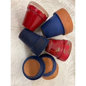 May include: A collection of small, painted terracotta flower pots. The pots are in shades of red and blue, with some showing signs of wear. The rims and saucers are visible, showcasing the natural terracotta color. Ideal for small plants or succulents.