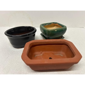 May include: Three small ceramic bonsai pots. One is black and oval-shaped, another is green and square with a matching saucer, and the third is a rectangular terracotta-colored pot.