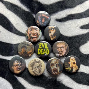 May include: A collection of ten round pinback buttons featuring images from the horror film "Evil Dead." The buttons display various character portraits and the film's title in yellow text. The buttons are arranged on a black and white zebra-striped surface.