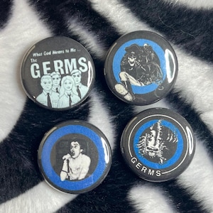 May include: Four black and white buttons with blue accents. One button reads "What God Means to Me... The GERMS" with a band photo. Other buttons feature illustrations and the band name. The buttons are on a black and white zebra print background.