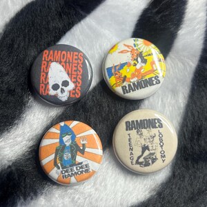 May include: Four round pinback buttons with graphics and text. One button features a skull and the word "Ramones". Another has a cartoon image and the word "Ramones". A third button has a cartoon image and the words "Dee Dee Ramone". The last button has the words "Ramones Teenage Lobotomy".
