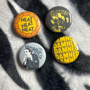 May include: Four round pinback buttons with different designs. One button has the text "NEAT NEAT NEAT" in yellow on an orange web design. Another has a black and white image of a band. The other two buttons have the text "THE DAMNED".