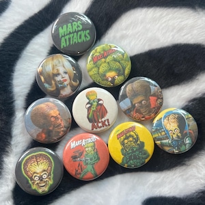 May include: A collection of nine circular pin-back buttons featuring various Mars Attacks character illustrations and the text Mars Attacks and ACK!. The buttons display diverse colors including black, yellow, orange, white, and green with detailed imagery.