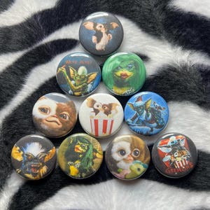 May include: A collection of round pinback buttons featuring various Gremlins characters. The buttons showcase colorful illustrations of the creatures, with a variety of backgrounds and designs. The buttons are arranged on a black and white zebra-striped surface.