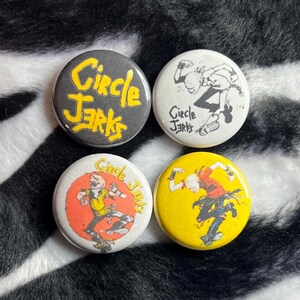 May include: Four round buttons with the text "Circle Jerks". The buttons have different designs, including a black button with yellow text, and buttons with illustrations of a person.