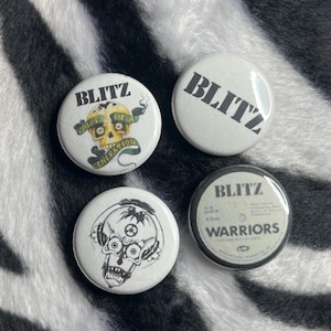 May include: Four white round buttons with the word "BLITZ" in bold black letters. One button features a skull with headphones, another has a skull with text, and a third has the words "WARRIORS". The buttons are on a black and white zebra-striped background.