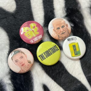 May include: A set of four circular pin-back buttons featuring imagery from The Substance. One button is pink with two faces, one shows a person, one is lime green with the text THE SUBSTANCE, and one is white with a yellow vial labeled ACTIVATOR.