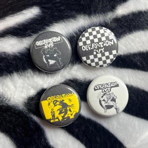 May include: Four round buttons with the text "OPERATION IVY". Two buttons are black and white, one with a checkered pattern. The other two buttons are black and yellow, and black and white with a graphic.