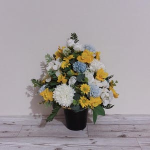 May include: A colourful floral arrangement in a black pot. The bouquet includes yellow, white, and blue flowers, with green leaves. The flowers are arranged densely, creating a cheerful and decorative display for a home.
