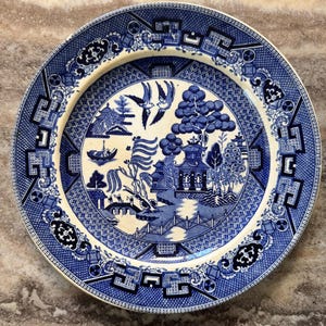 May include: A blue and white ceramic plate with a Willow pattern design. The plate features a central landscape scene with a bridge, pagoda, and birds, surrounded by a detailed border. The plate is round and has a cream-colored rim.