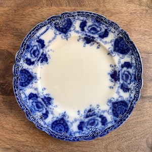 May include: A round, antique white plate with a blue floral design. The plate features a scalloped edge and a repeating pattern of roses and heart-shaped motifs. The plate's design is a classic, vintage style.