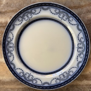 Puede incluir: Un plato de cerámica blanco con un borde decorativo azul y gris. El borde presenta un patrón repetitivo de diseños florales y de volutas. El plato tiene un centro color crema, probablemente para servir alimentos.