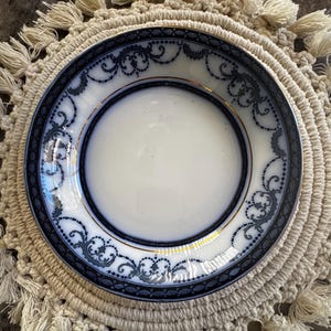 Puede incluir: Un plato de porcelana blanca con un borde decorativo azul marino y dorado. El plato presenta un diseño ornamentado y está colocado sobre un salvamanteles de macramé beige con flecos.