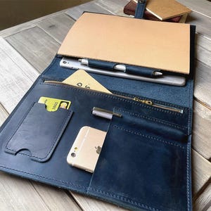 May include: A dark blue leather portfolio with a zipper closure. The portfolio has multiple pockets and slots for holding a phone, pens, and cards. The portfolio is open and the interior is visible.