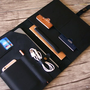 May include: A black leather organizer folio featuring multiple interior pockets and slots. It contains a smartphone, a white charging cable, credit cards, a notebook, and a passport. The folio is secured with a strap and a metal snap button closure.