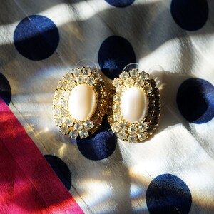 May include: A pair of gold oval-shaped earrings with white pearl-like stones set in a cluster of clear rhinestones.