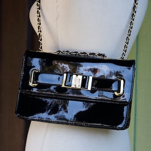 May include: A black patent leather handbag with a gold chain strap and a gold buckle closure. The bag has a flap with a bow detail.