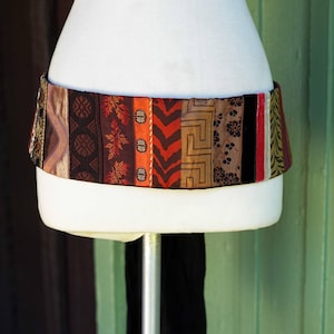 May include: A wide, patterned belt with various fabric panels in shades of brown, orange, and red. The belt is displayed on a white mannequin, showcasing its design and texture. The belt's design includes geometric and floral patterns.
