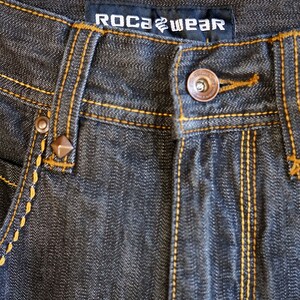 Peut inclure: Gros plan sur un jean en denim gris fonc&eacute; avec des coutures jaunes. Le jean porte une &eacute;tiquette de marque qui indique "ROCAWEAR".