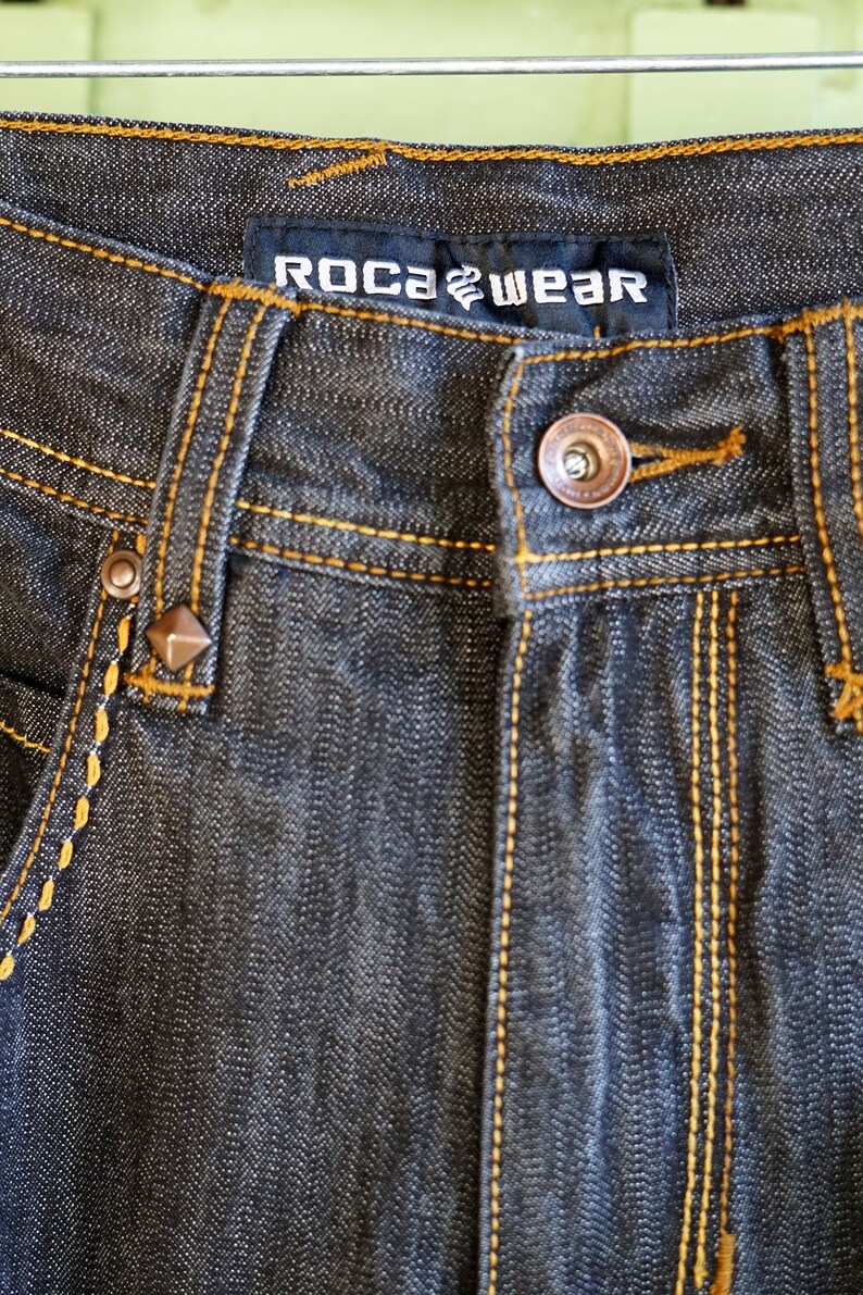 Peut inclure: Gros plan sur un jean en denim gris fonc&eacute; avec des coutures jaunes. Le jean porte une &eacute;tiquette de marque qui indique "ROCAWEAR".