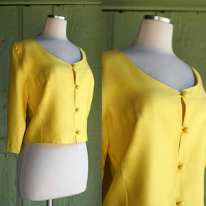 May include: A bright yellow, button-down jacket with a V-neckline and three-quarter sleeves. The jacket is displayed on a mannequin, showcasing its tailored fit and classic design. The buttons are the same yellow color as the jacket.