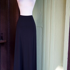 May include: A black maxi skirt displayed on a mannequin. The skirt has a high waist and a flowing silhouette. The hemline features a white border with a dotted design. The mannequin is set against a backdrop of a wooden door.