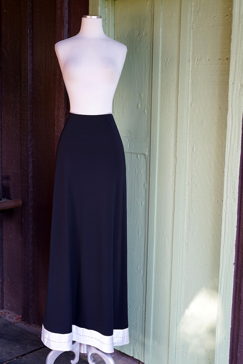 May include: A black maxi skirt displayed on a mannequin. The skirt has a high waist and a flowing silhouette. The hemline features a white border with a dotted design. The mannequin is set against a backdrop of a wooden door.