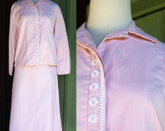 Könnte beinhalten: Ein hellrosa zweiteiliger Rockanzug. Die langärmlige Jacke hat einen Kragen und weiße Gänseblümchenbesatz auf der Vorderseite. Der Rock ist knielang. Der Anzug wird auf einer Schaufensterpuppe präsentiert.